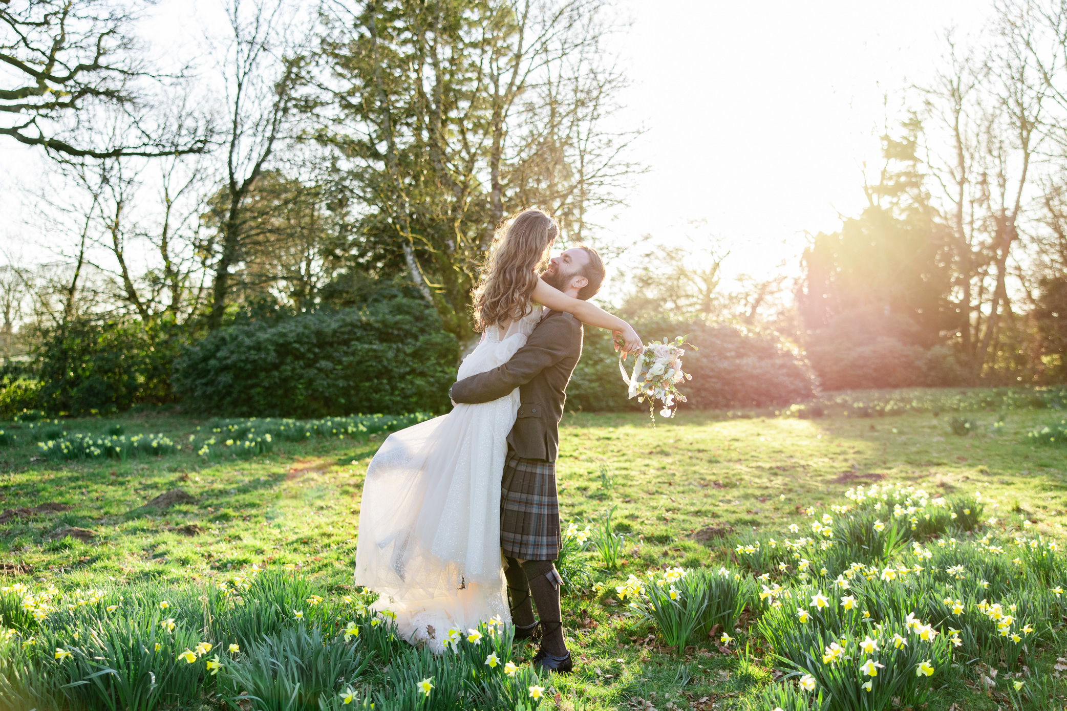 kilt wearing groom lifts bride in glowy sunlight