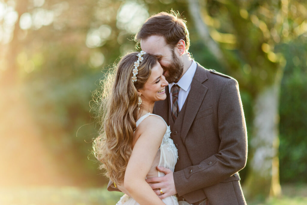 kilt wearing groom smiles and snuggles bride in glowy sunlight