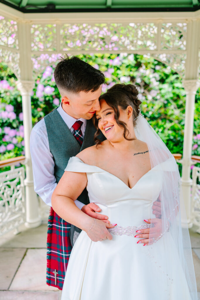 Bride and groom cuddling beneath the ornate gazebo at Kilmardinny House wedding venue near Glasgow