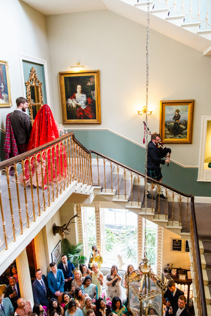 Bride and groom descend the grand staircase at Melville Castle while a bagpiper leads guests during their wedding celebration