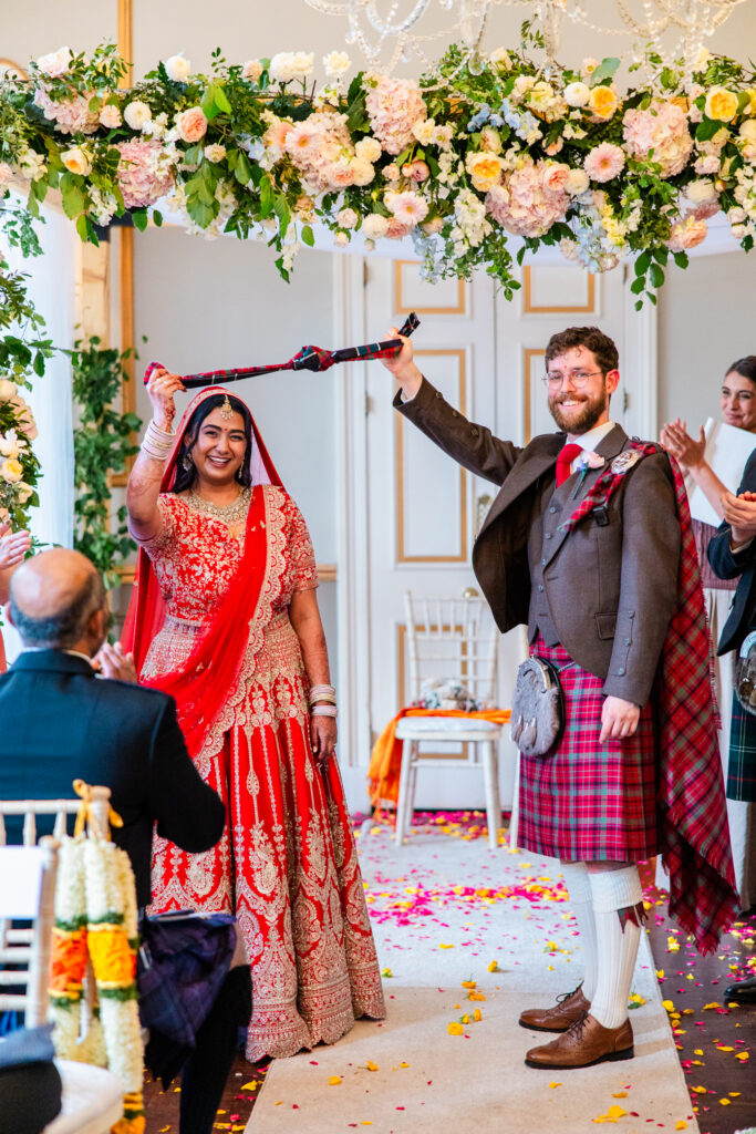 Bride and groom holding tartan ribbon during a Scottish and Indian wedding ceremony at Melville Castle beneath a floral arch