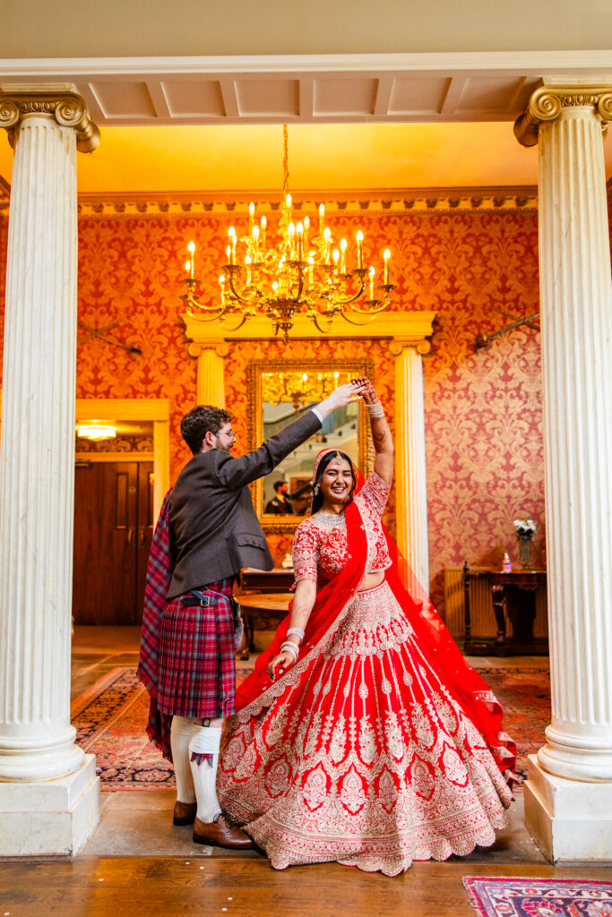 Bride in a red lehenga twirling with her groom in a Scottish kilt inside Melville Castle