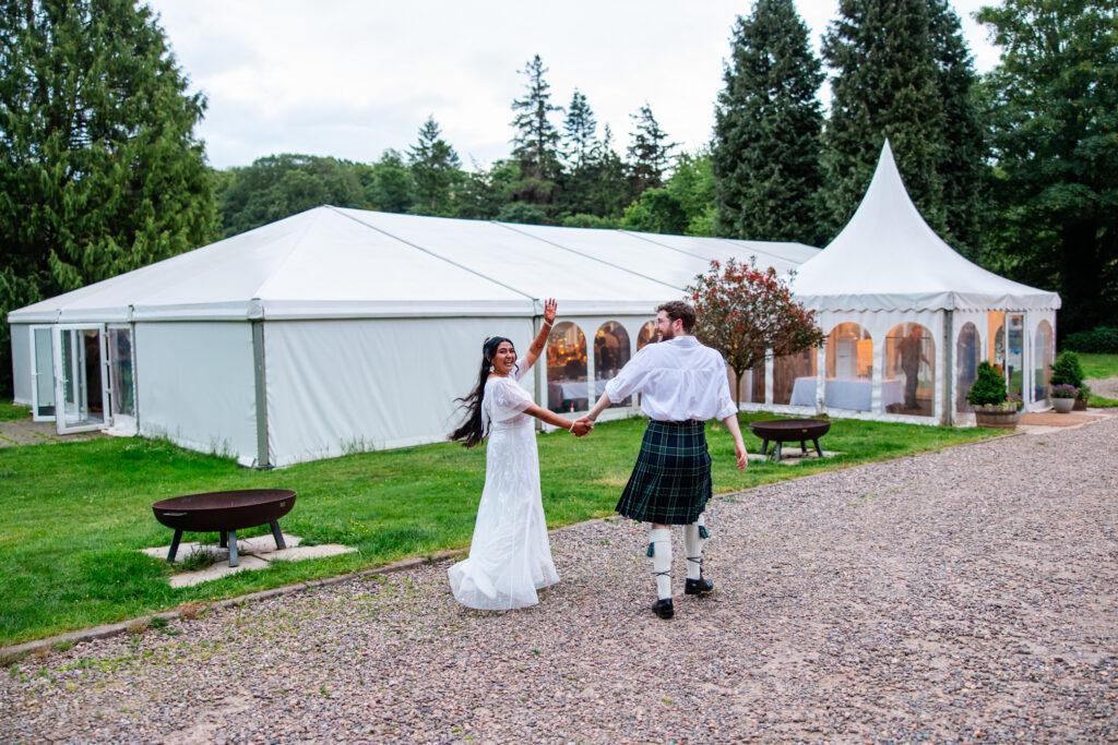 Bride waving while walking hand in hand with her groom past the wedding marquee at Melville Castle wedding venue in Scotland