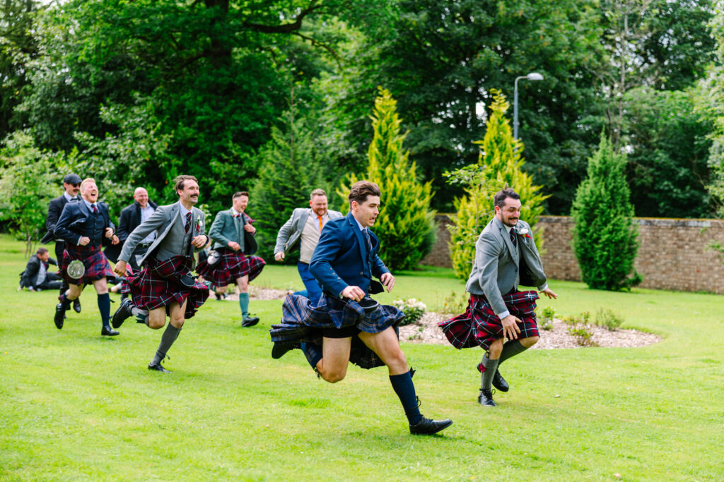 Groomsmen sprinting in kilts during an outdoor wedding race at Kilmardinny House Glasgow