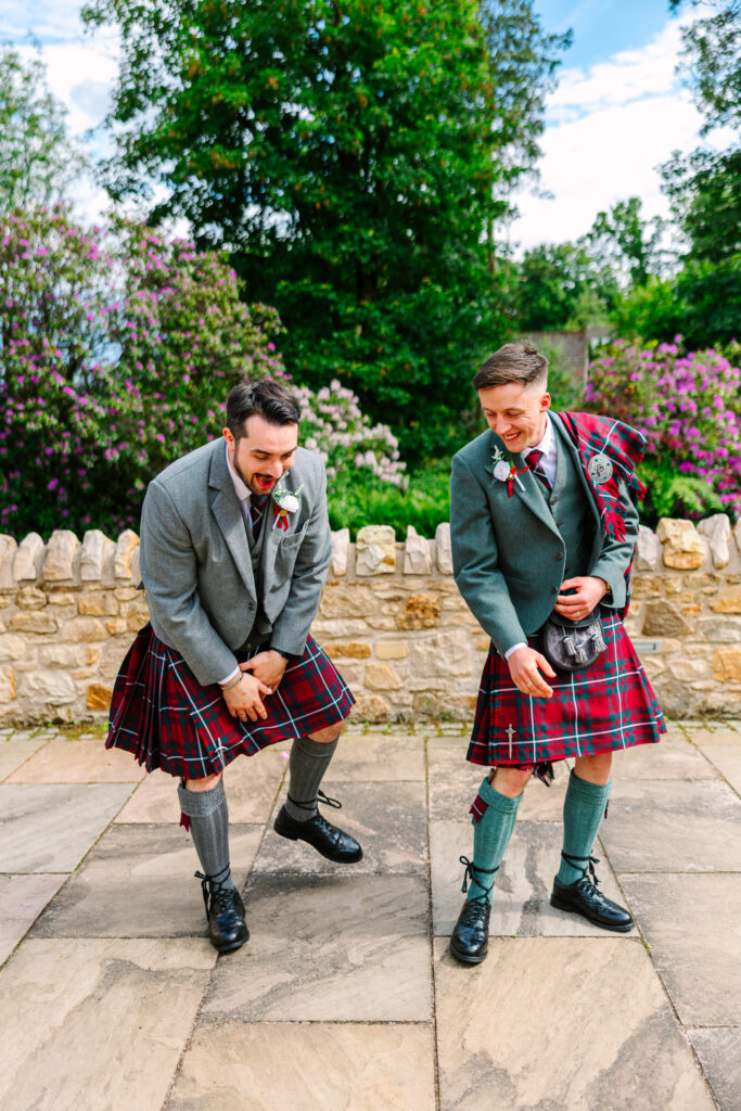 Two groomsmen sharing a laugh in kilts during wedding portraits in the gardens at Kilmardinny House