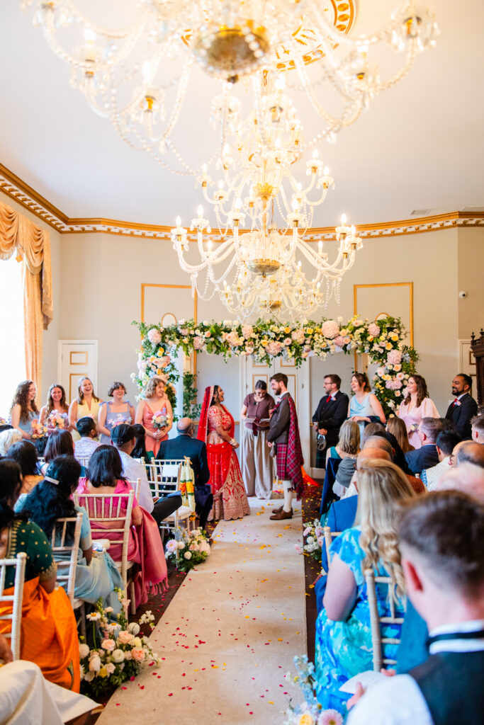 Wide view of a Scottish and Indian wedding ceremony at Melville Castle with guests, floral arch and crystal chandelier