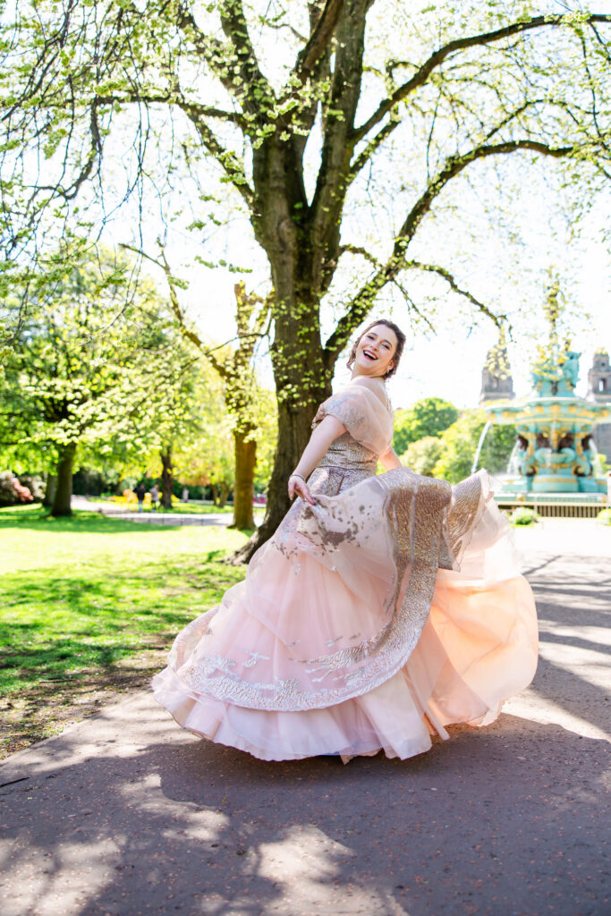 A woman laughing as she twirls in a flowing blush pink and silver gown in Princes Street Gardens, Edinburgh, with Ross Fountain behind her