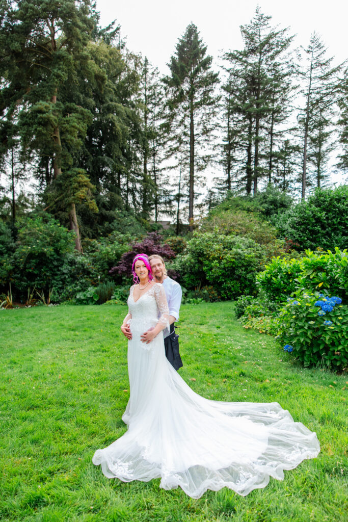 Bride standing in the gardens at Balgeddie House, surrounded by lush greenery and trees, with her long wedding dress flowing across the grass