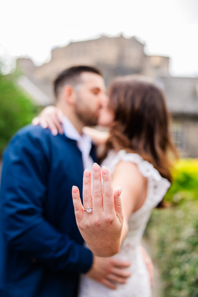 Close-up of engagement ring with the couple kissing and Edinburgh Castle softly blurred behind