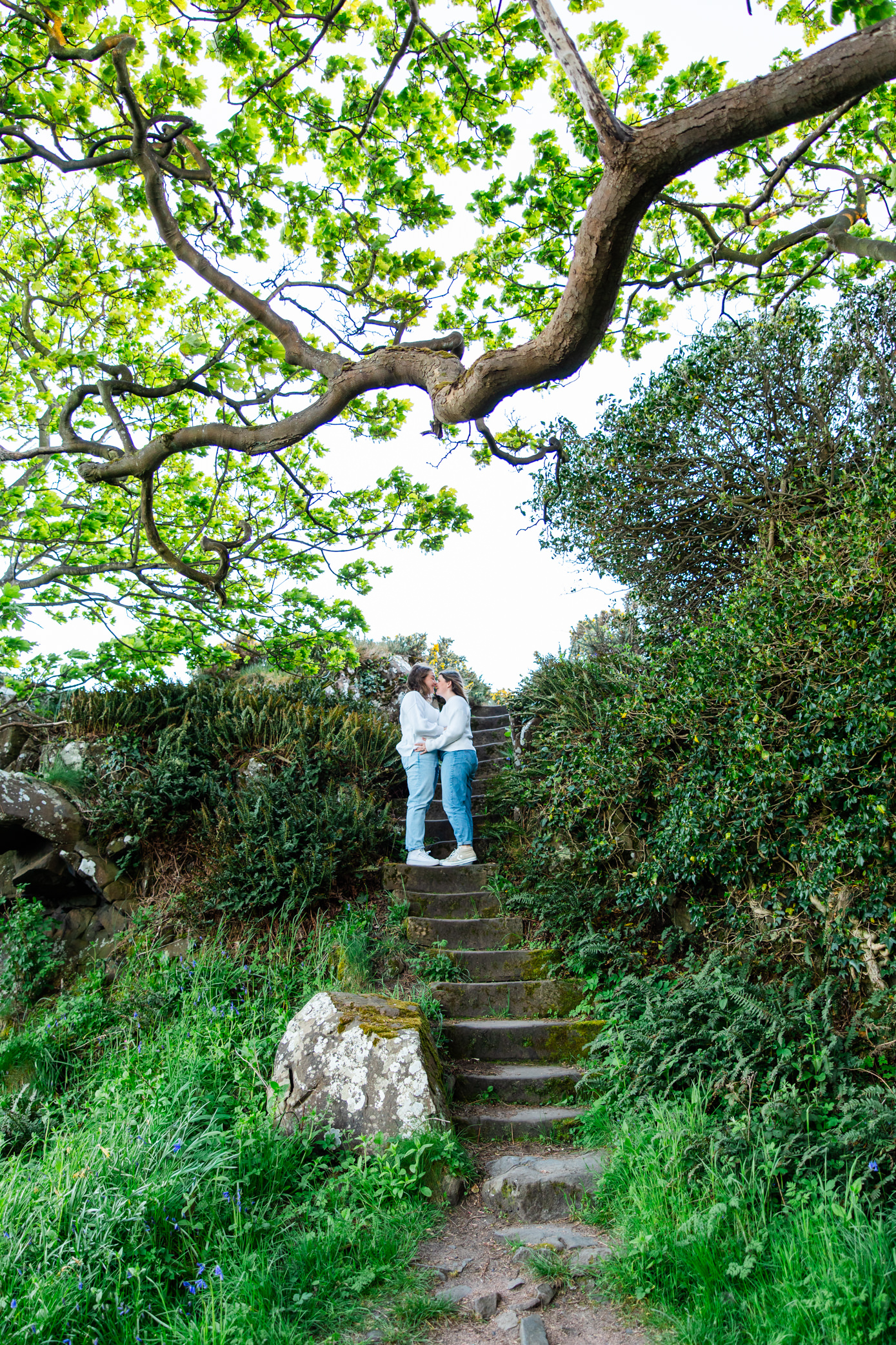 Queer couple standing together beneath a large tree on woodland steps at Downing Point in Scotland