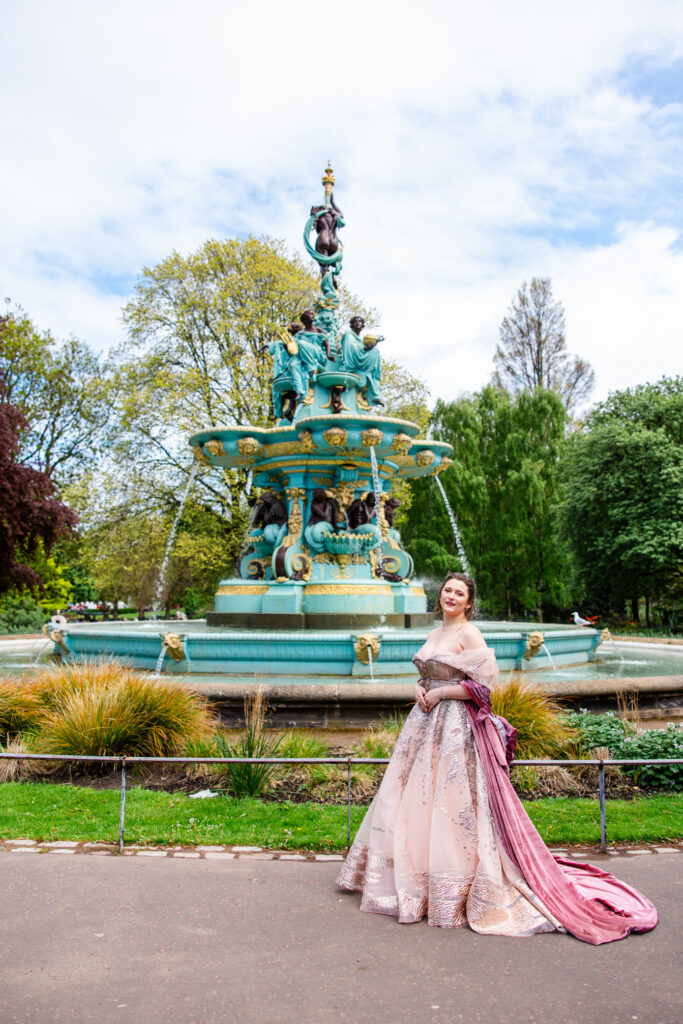 Woman in a flowing blush pink ball gown standing beside Ross Fountain with Edinburgh Castle in the background