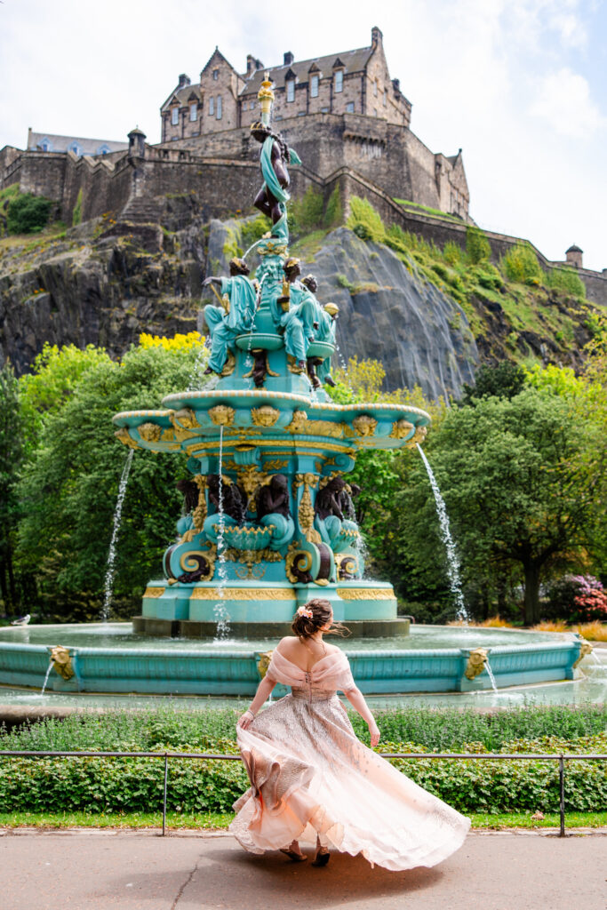 Woman in a pink medieval ball gown posing beneath Ross Fountain with Edinburgh Castle rising behind her