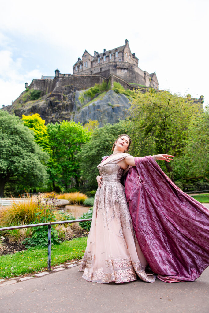 Woman wearing a pink medieval-style gown and dramatic burgundy cloak overlooking Edinburgh with the castle behind her